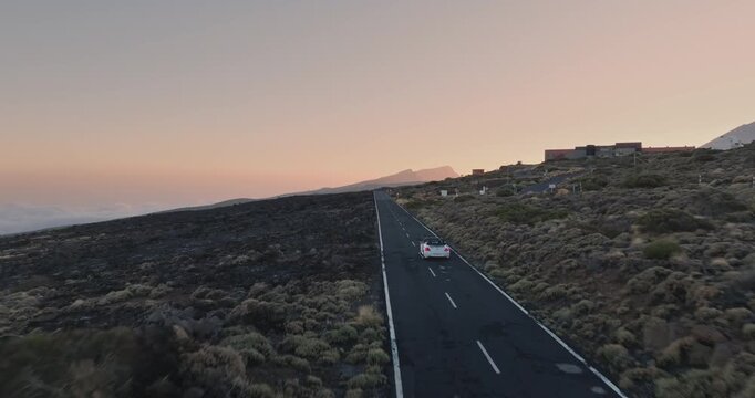 Car driving on a long desert road at sunset. Surrounded by sparse mountain vegetation and a distant observatory. With colorful sky and cloud layer visible on the horizon. Aerial tracking drone flight