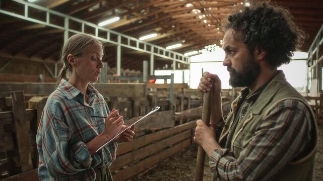 Female farm manager reviewing checklist on clipboard and explaining daily tasks to bearded male worker holding pitchfork handle inside rustic livestock barn