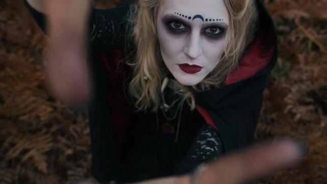Top view shows pale faced woman wearing black hooded cloak reaching upward toward viewer with blurred hand in foreground, dark occult makeup, intense expression, and moody forest backdrop