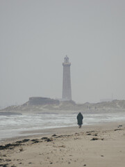 Lighthouse emerging through coastal mist as a lone figure walks along a Danish beach beside rough sea. Ideal for travel, solitude, maritime heritage and atmospheric landscape themes. © Pavel