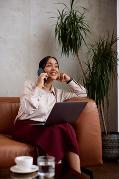 Smiling businesswoman talking on mobile phone while using laptop at hotel reception area.