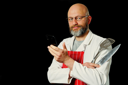 Bald man with glasses and beard stands confidently. He wears white coat and red striped apron. Holds spatula in hand. Smiles at camera. Black background highlights his figure