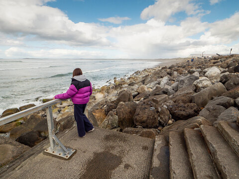 Child in a purple jacket stands on stone steps by a rocky shoreline. Waves crash against large, wet boulders under a cloudy sky. The child looks out at the ocean, Strandhill, Sligo, Ireland. Travel