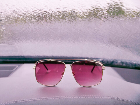 Pink tinted sunglasses rest on car dashboard. Raindrops visible on windshield behind. Car interior with red trim details. Outdoors view through wet glass. Reflections show trees and sky
