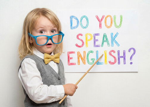 Cute little blonde European girl in a vest and bow tie wearing blue glasses. She acts as a teacher pointing at a Do You Speak English text. Educational concept for language kids.