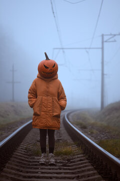 Pumpkin headed figure standing alone on railway tracks disappearing into dense fog. Surreal Halloween concept with abandoned railroad and eerie autumn atmosphere