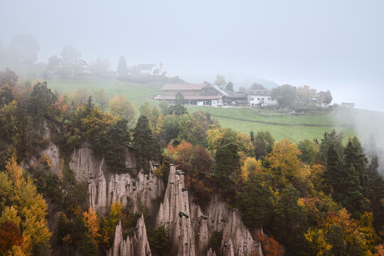 Renon Earth pyramids beneath a foggy hillside village surrounded by autumn forests in South Tyrol, Dolomites, Italy. Scenic landscape with unique rock formations and fall colors