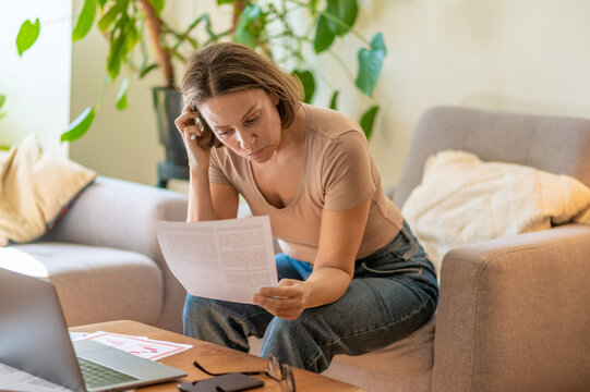 Stressed middle aged woman holding document near laptop