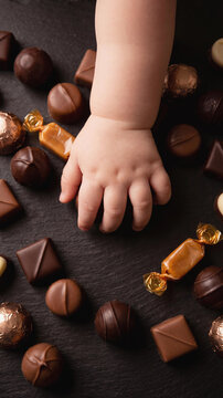 High angle shot of a sweet chubby baby hand reaching for a round chocolate praline among a large assortment of delicious milk and dark candies on a textured black slate background.