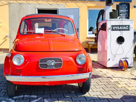 Front view of a vintage red Steyr-Puch 500 veteran car, based on the Fiat 500 design, parked on a paved lot next to a public car vacuum cleaner station.