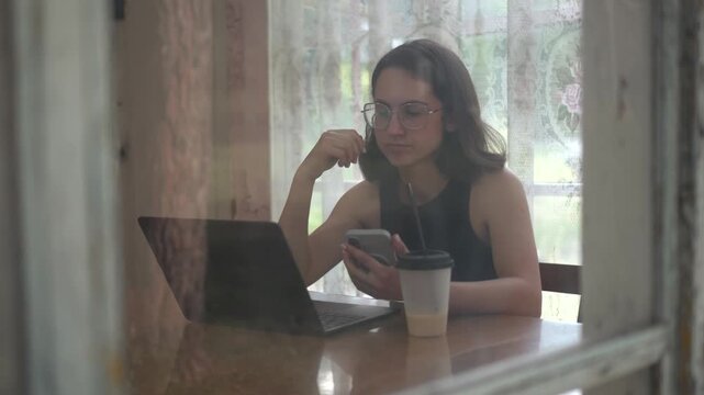 Young woman using laptop and smartphone while drinking coffee on old glass veranda