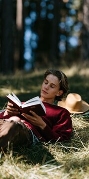 Reclining young reader on grassy forest floor with a hat nearby reading a novel for calm background and outdoor lifestyle banner design