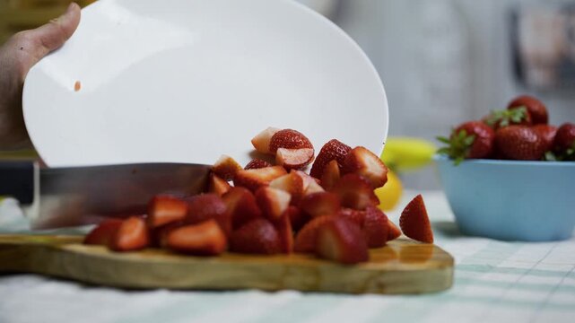 Person using knife to move sliced strawberries onto cutting board in kitchen, closeup shot
