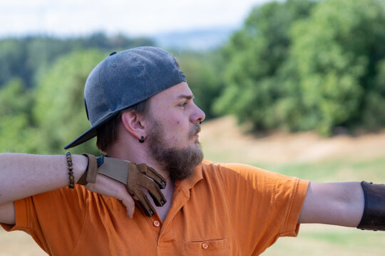Young male bearded adult with orange shirt, cap, arm guard and a leather glove is standing just after releasing the arrow from the bow to hit his target outside .