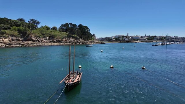 DOUARNENEZ, FRANCE &ndash; July 4, 2026: Calm coastal harbor with a traditional sailboat and mooring buoys in Brittany, France. Rocky shores and the town waterfront appear in the distance.