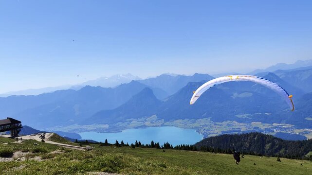 A paraglider prepares for takeoff on a grassy mountain summit overlooking a vast blue lake and green hills under a clear sky.