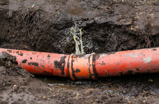 A damaged orange underground pipe leaking water and mud in an excavated trench, illustrating plumbing accidents, infrastructure damage, and pipe repair work
