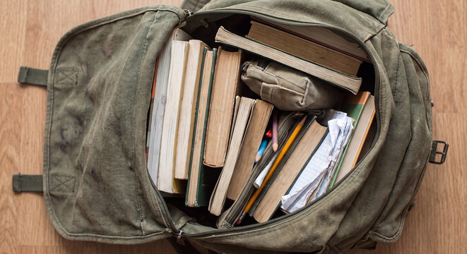 An overhead view of an open vintage green backpack packed with various books lying on a rustic wooden surface in a cozy learning concept