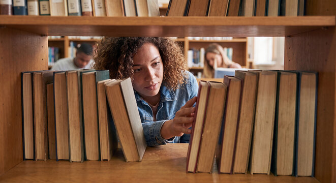 Young woman looking through a row of old books on a wooden bookshelf inside a quiet library or bookshop