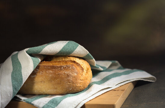 Rustic crusty homemade artisan bread loaf under linen cloth on wooden cutting board with dark background and copy space.