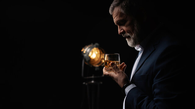 Handsome businessman in stylish suit holding glass whiskey on dark