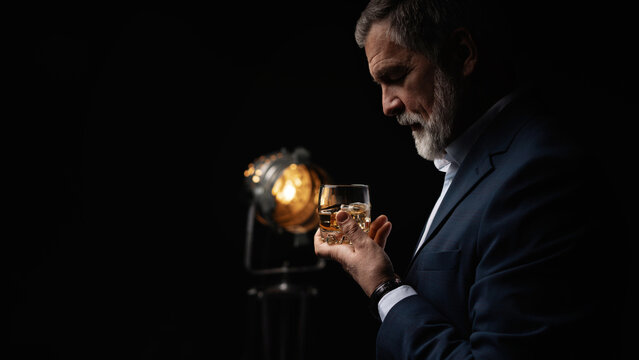 Handsome businessman in stylish suit holding glass whiskey on dark