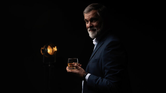 Handsome businessman in stylish suit holding glass whiskey on dark