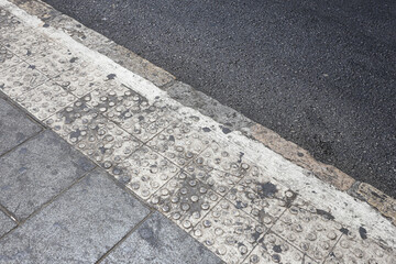 Fototapeta premium Tactile paving runs beside asphalt road. Raised circles cover worn concrete tiles. White painted edge follows the curb. Dark stains mark the walking surface. Street textures form an urban background.