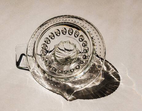 Top view of a vintage clear glass citrus juicer illuminated by direct sunlight, casting intricate reflections and shadows on a neutral surface.