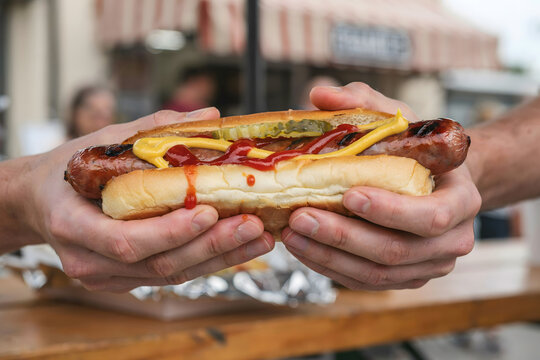 A person holding a grilled hot dog in a soft bun with mustard, ketchup, and relish, with steam rising from the warm sausage at a food stand