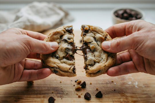 Close-up of hands breaking a warm chocolate chip cookie in half on a wooden board, revealing a gooey melted chocolate center and dark crumbs