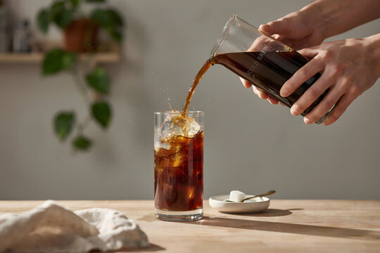 Pouring cold brew coffee from a glass carafe into a tall glass filled with ice cubes on a wooden table with sugar cubes on a small plate