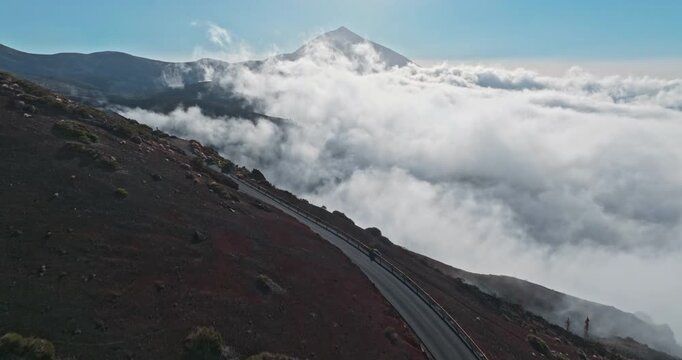Teide National Park landscape showing a winding mountain road on volcanic terrain, extending above a blanket of white clouds with the peak of the Teide volcano rising in the background