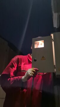 Electrician inspecting junction box, technician in red uniform checking high voltage outdoor fuse cabinet at night. Low angle handheld camera shot.