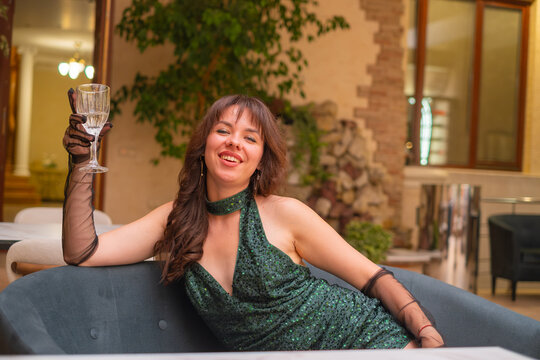 Woman in a green dress is sitting on a couch in a lounge and holding a glass while smiling