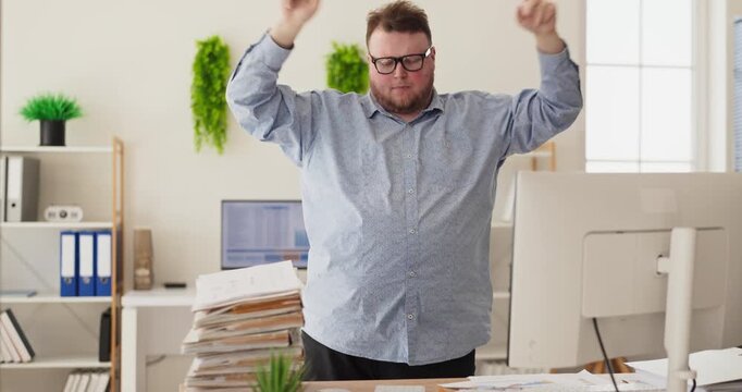 Overweight man dancing in office near computer. Happy worker celebrates success and fun at work amid paperwork in a business office. Concept of analysis celebration workplace motivation.