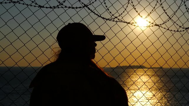Woman looking at sunset behind barbed wire fence. Woman standing behind barbed wire fence, silhouetted against golden sunset over ocean, with sense of solitude and longing for freedom