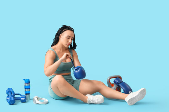 Female bodybuilder with sports equipment putting boxing glove on blue background