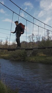 Hiker with a backpack walking across a narrow metal suspension bridge over a flowing mountain river, surrounded by trees and mountains.