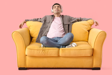 Fototapeta premium Handsome young man in headphones sitting on yellow sofa against pink background