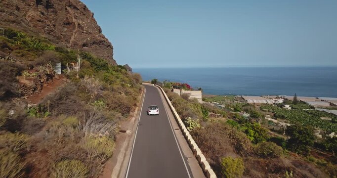 White small car driving along scenic coastal road in Tenerife, Canary Islands, steep rocky mountain cliff on one side and the vast blue ocean stretching to the horizon on the other side. Aerial shot