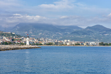 Obraz premium Scenic view of Alanya, Turkey featuring a lighthouse along the coastline with mountains and cityscape in the background under a cloudy sky