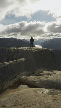 Back view of a hiker with a backpack standing on a rocky cliff edge, admiring the panoramic view of a deep mountain lake surrounded by steep terrain.