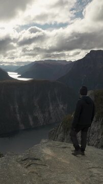 Back view of a hiker standing on a high rocky cliff, observing the expansive mountain landscape and a distant lake under a cloudy sky.