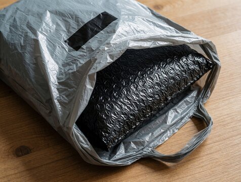Opened gray plastic shipping bag lies on wooden table. Black bubble wrap protects parcel inside mailer envelope. Online shop customer unboxing delivery package at home. E-commerce logistics service