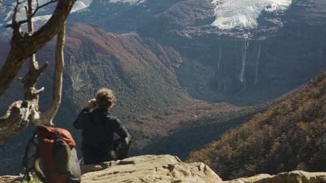 Hiker with a backpack sitting on a rocky ledge, overlooking the scenic Casta&ntilde;o Overa glacier and cascading waterfalls in Nahuel Huapi National Park, Argentina.