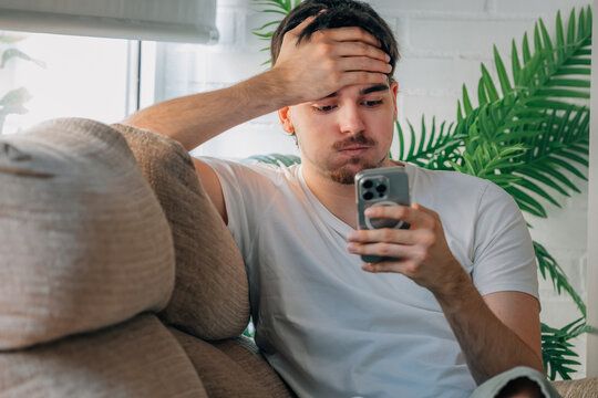 Worried young Caucasian man sitting on a sofa at home, holding smartphone with stressed expression and hand on forehead, reading bad news or shocking message, anxiety and stress concept
