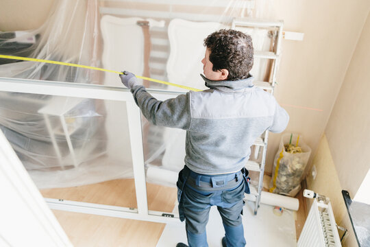A full-length portrait of a worker measuring a window frame indoors with a tape measure before installation.