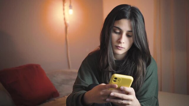 Young Latina woman in her bedroom texting on her smartphone while blushing with a shy smile. Authentic lifestyle, private communication, emotional moment at home.
