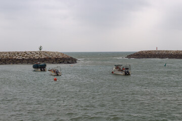 Boats navigate a coastal harbor during an overcast day near rocky jetties © Toyagrafia